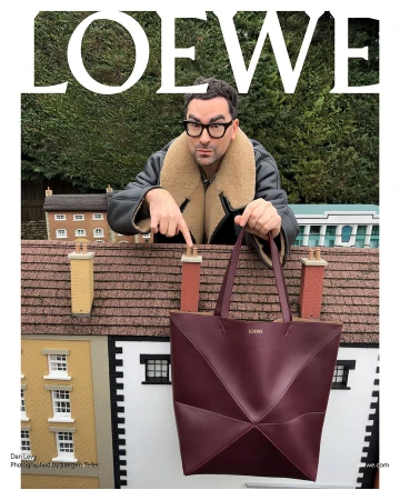Dan Levy models a Loewe tote bag against a miniature town backdrop, wearing a shearling jacket and glasses in an outdoor setting.