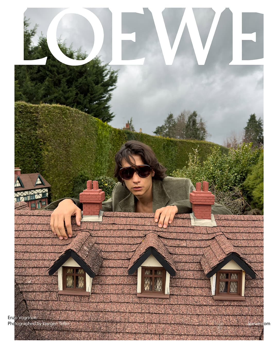 Man in sunglasses peers over the roof of a miniature house outdoors against a green hedge and cloudy sky, Loewe text at top.