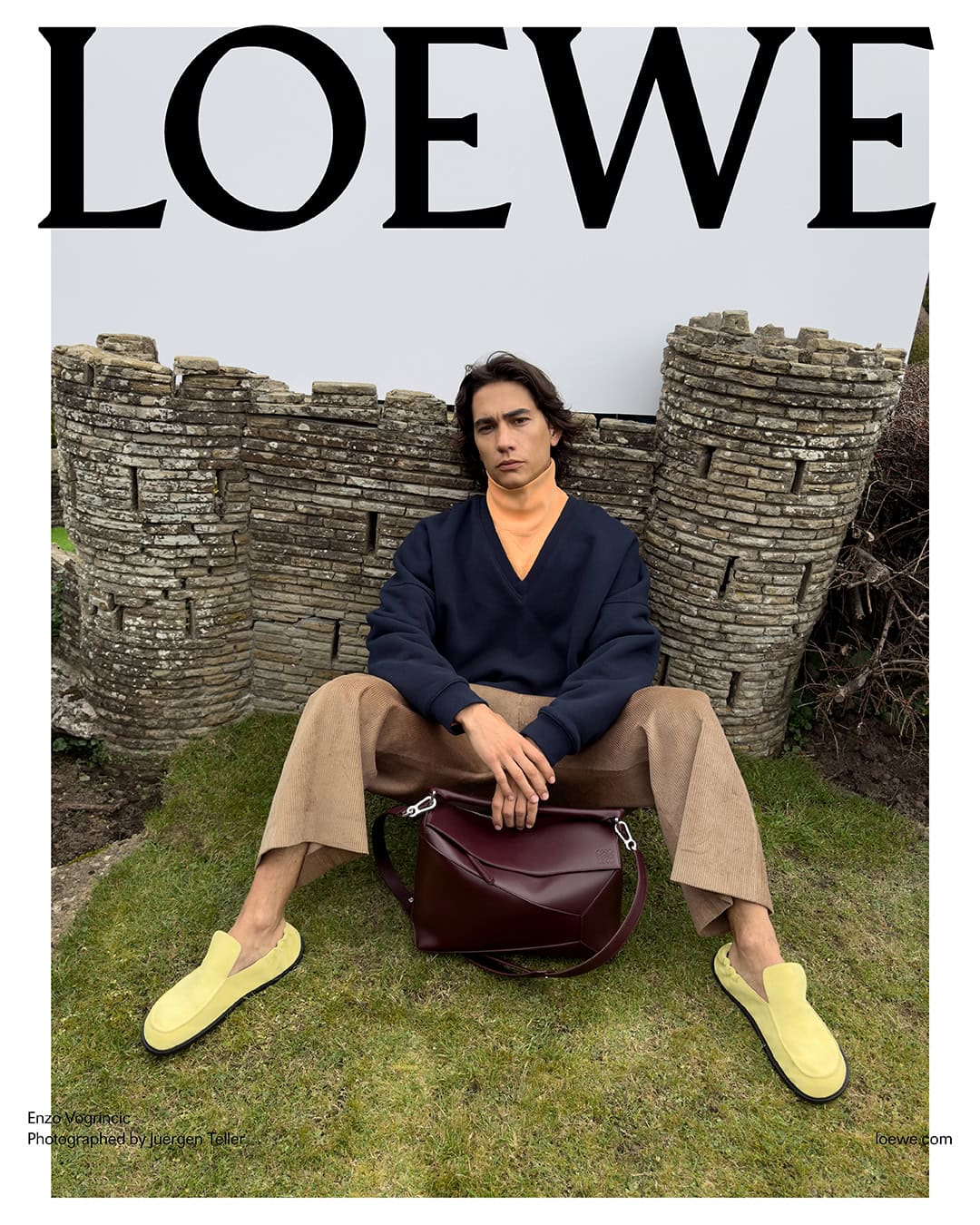 Male fashion model poses on green grass wearing a sweater, trousers and loafers with a satchel against a stone wall backdrop.