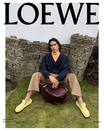 Male fashion model poses on green grass wearing a sweater, trousers and loafers with a satchel against a stone wall backdrop.