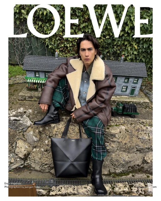 A man models a Loewe jacket and plaid pants while sitting on a stone wall with a Loewe handbag, greenery, and a toy village in the background.