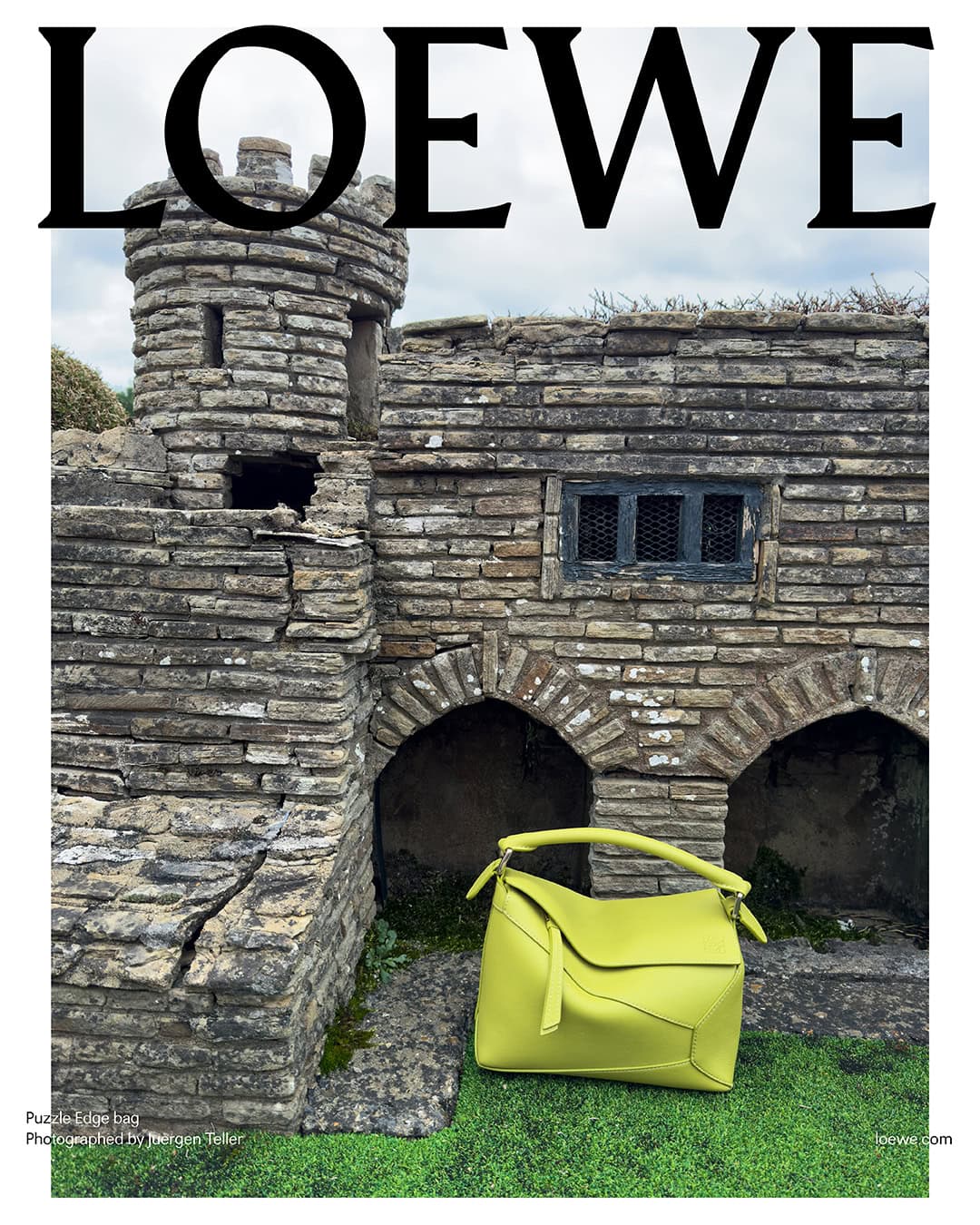 A bright yellow handbag is positioned next to a miniature stone building under a cloudy sky, featuring the bold 'LOEWE' logo.