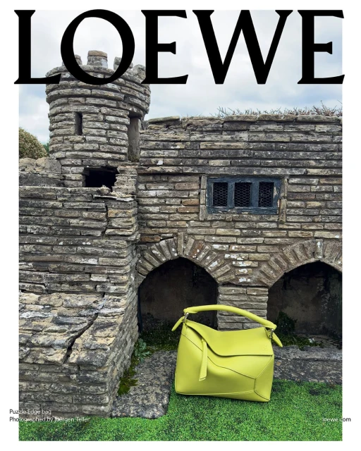 A bright yellow handbag is positioned next to a miniature stone building under a cloudy sky, featuring the bold 'LOEWE' logo.