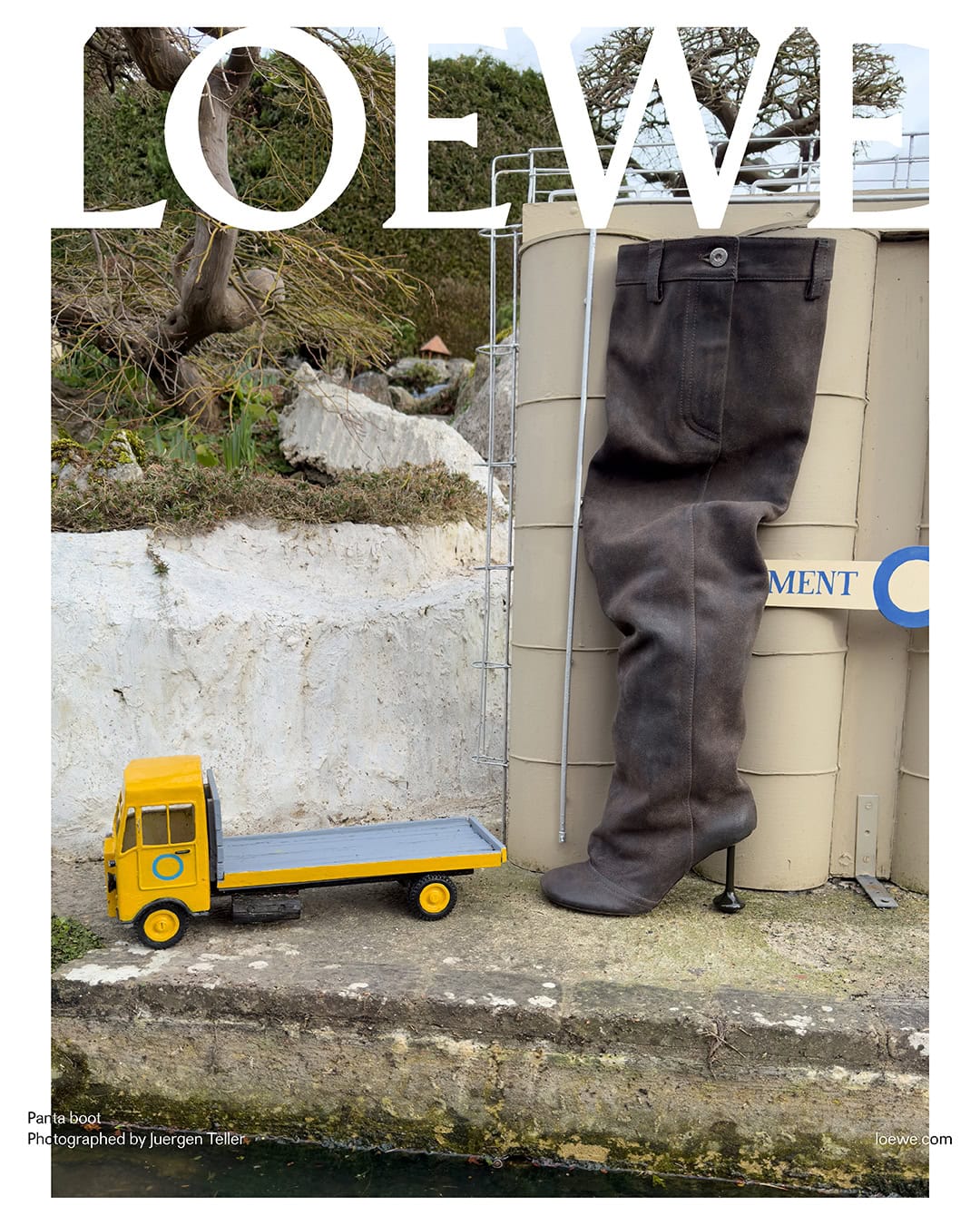 Fashion editorial featuring a tall boot alongside a miniature yellow truck, set against an outdoor industrial backdrop with the Loewe logo.