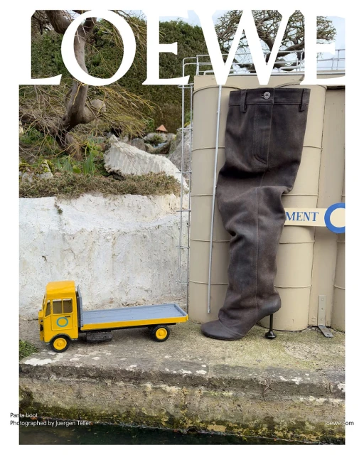 Fashion editorial featuring a tall boot alongside a miniature yellow truck, set against an outdoor industrial backdrop with the Loewe logo.