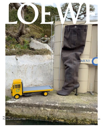 Fashion editorial featuring a tall boot alongside a miniature yellow truck, set against an outdoor industrial backdrop with the Loewe logo.
