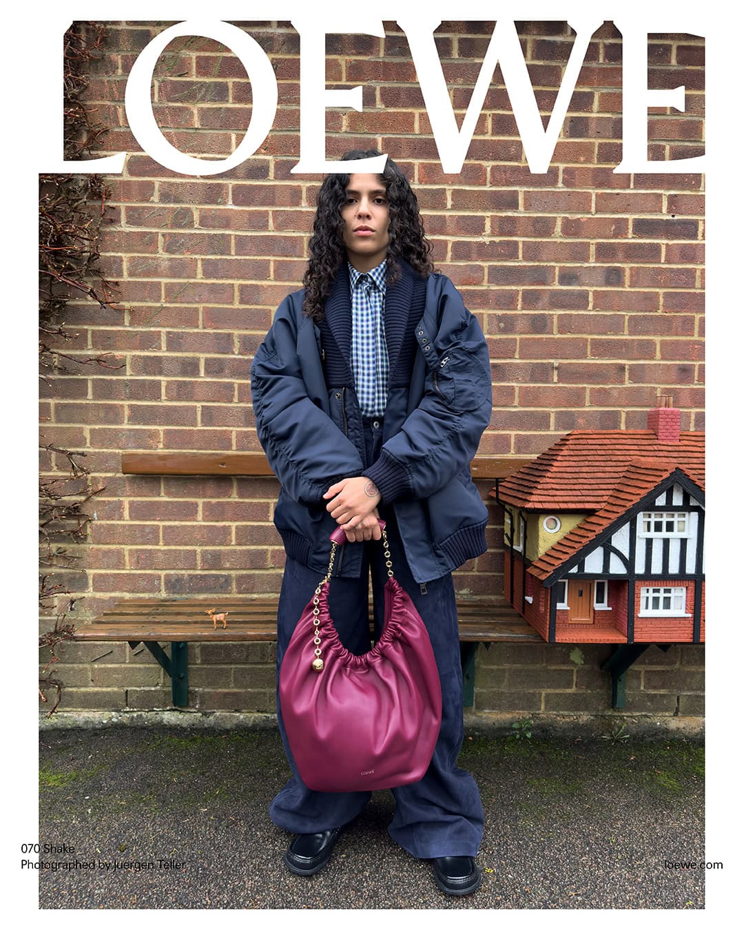 A fashion editorial shot of a person holding a maroon Loewe bag in an outdoor setting, featuring a brick wall and a decorative birdhouse.