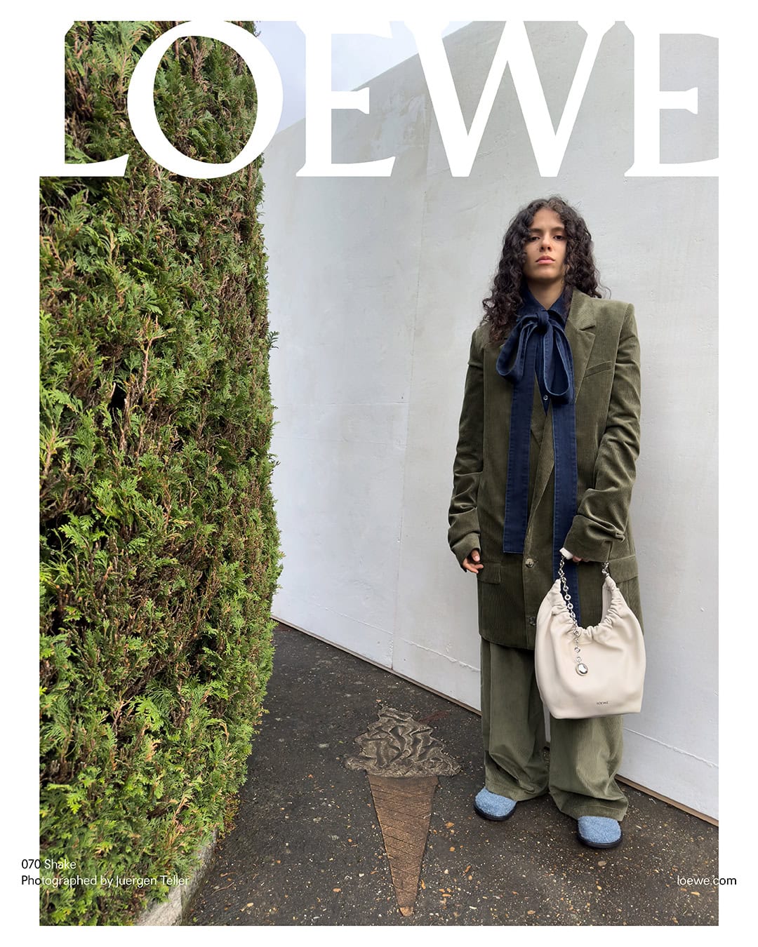 Fashion editorial featuring a woman in an olive-green suit carrying a white Loewe bucket bag, posing against a textured wall and hedge.