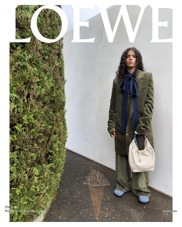 Fashion editorial featuring a woman in an olive-green suit carrying a white Loewe bucket bag, posing against a textured wall and hedge.
