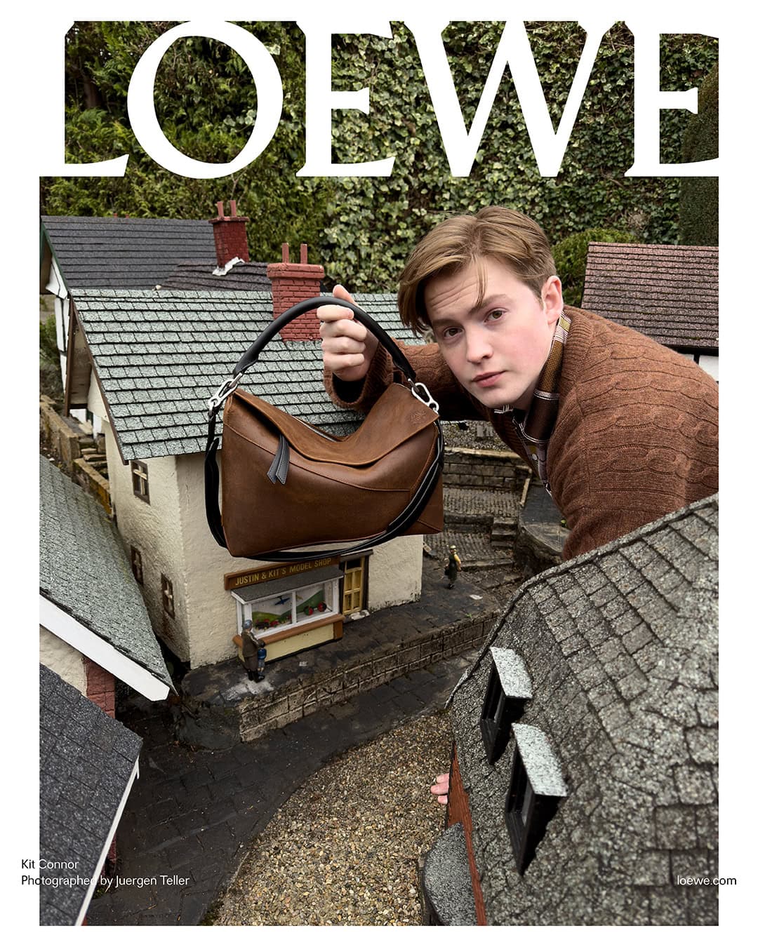 Fashion editorial shot of a male model holding a brown leather handbag standing on a miniature building with greenery background and LOEWE text.