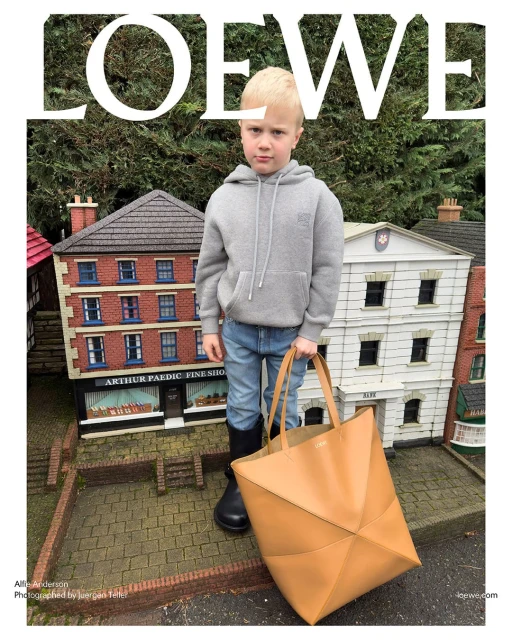 Child model wearing a gray hoodie, jeans, and black boots, stands with a tan leather bag in front of miniature buildings and greenery.