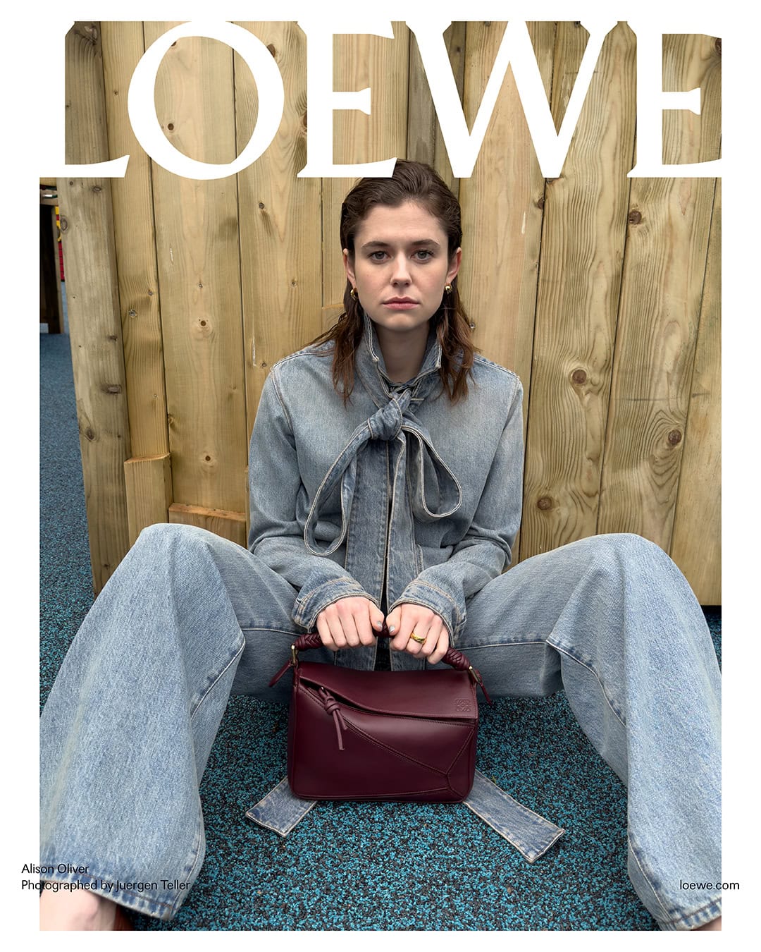 A woman sits in front of a wooden wall, wearing a denim outfit and holding a dark red handbag with the Loewe brand displayed on top.