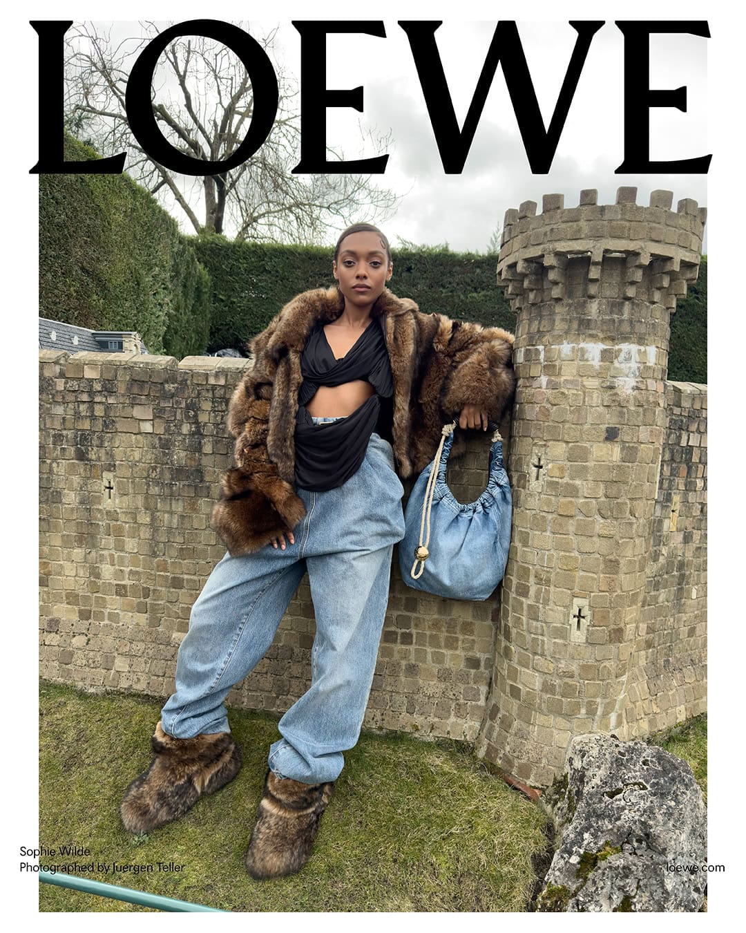 A woman in a fur coat and jeans poses near a stone structure in an outdoor fashion shot for Loewe, showcasing a unique bag and footwear.