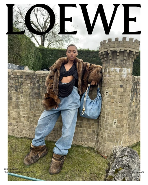 A woman in a fur coat and jeans poses near a stone structure in an outdoor fashion shot for Loewe, showcasing a unique bag and footwear.