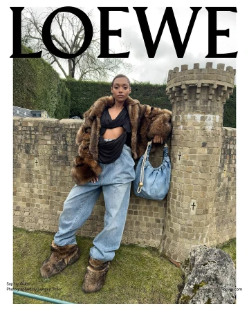 A woman in a fur coat and jeans poses near a stone structure in an outdoor fashion shot for Loewe, showcasing a unique bag and footwear.