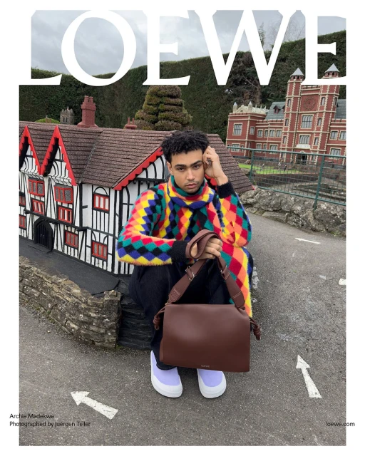 An adult male sits outdoors with a brown Loewe bag, wearing a colorful argyle sweater and light purple shoes, near miniature buildings.