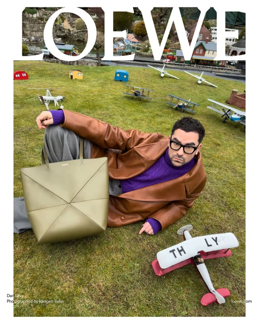 Man in brown leather jacket poses with Loewe bag in outdoor setting with model airplanes and miniature buildings. Fashion editorial shot.
