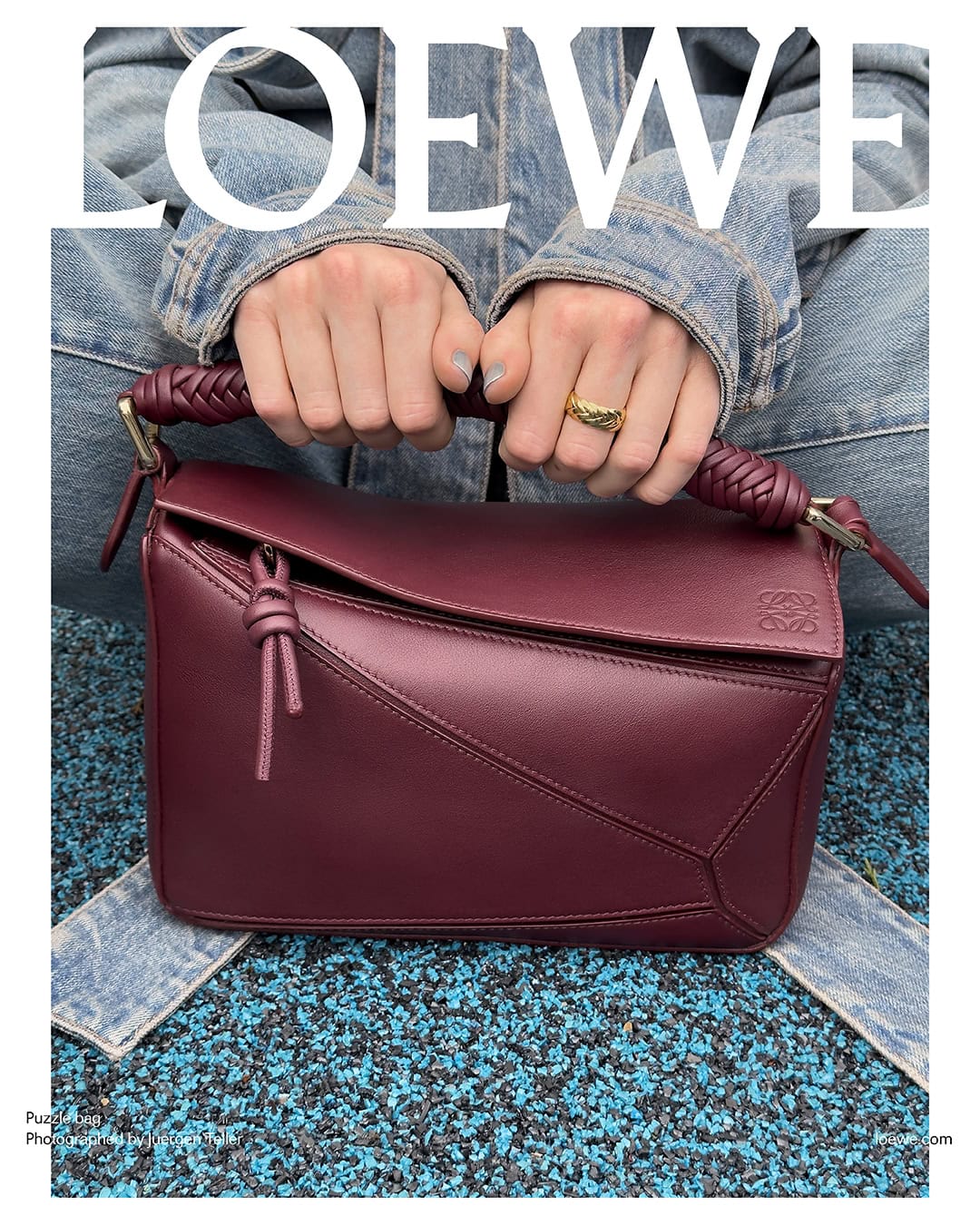Burgundy Loewe Puzzle bag is held by an adult female wearing a denim jacket and gold ring, against a blue and black speckled background.