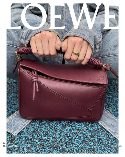 Burgundy Loewe Puzzle bag is held by an adult female wearing a denim jacket and gold ring, against a blue and black speckled background.