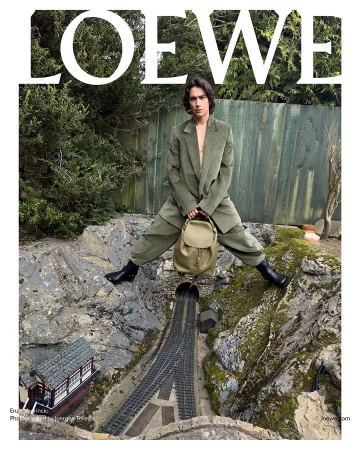 Fashion editorial shot of a slender adult man in an olive suit holding a backpack, posing near model train tracks and miniature buildings.