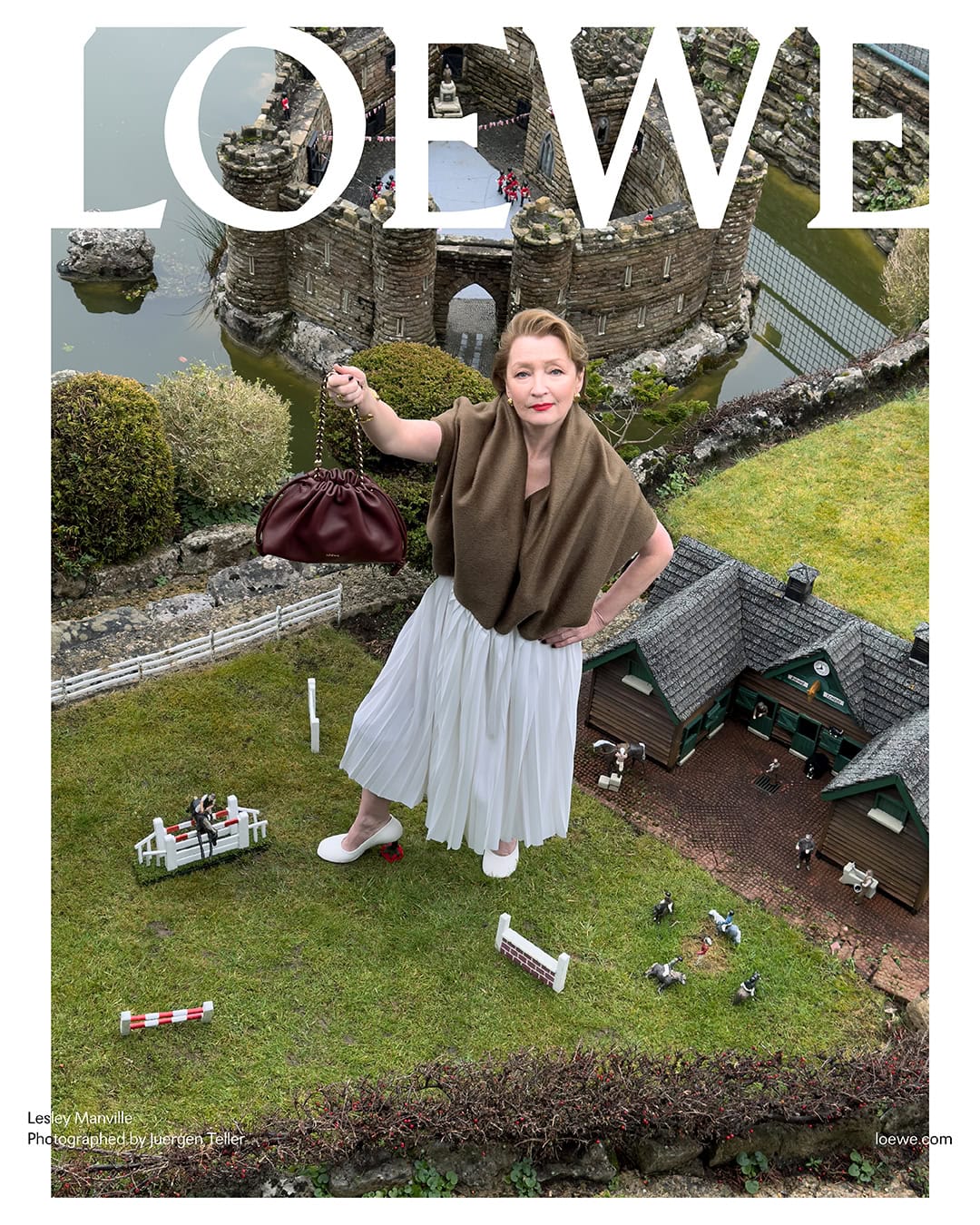 Fashion editorial image of a woman posing with a burgundy handbag against a miniature landscape with a castle and green grass.