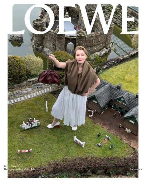 Fashion editorial image of a woman posing with a burgundy handbag against a miniature landscape with a castle and green grass.