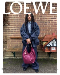 A person standing outdoors in front of a brick wall holding a magenta handbag, wearing a navy jacket and trousers.