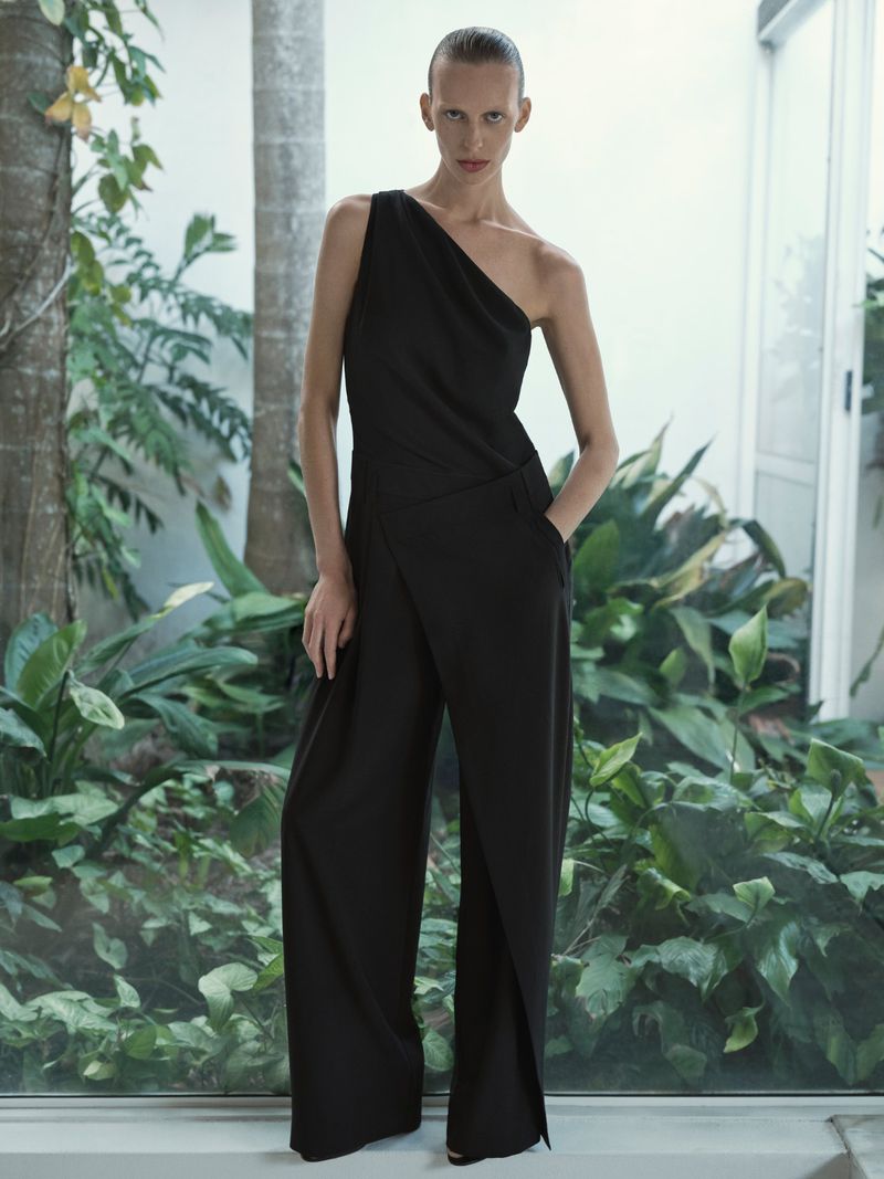 Fashion model poses in one-shoulder black jumpsuit against a backdrop of green plants and natural light, creating an elegant portrait.