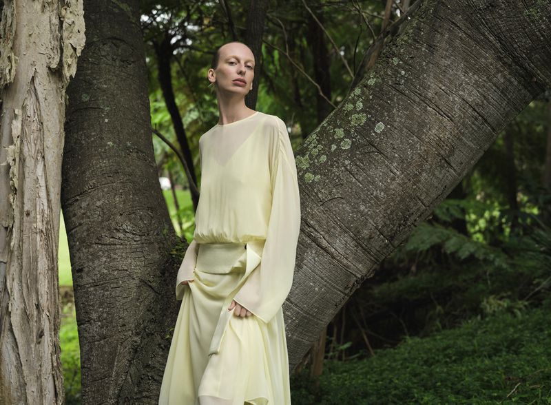 An adult female model in a pale yellow dress poses in an outdoor setting surrounded by trees and green vegetation for a fashion editorial.