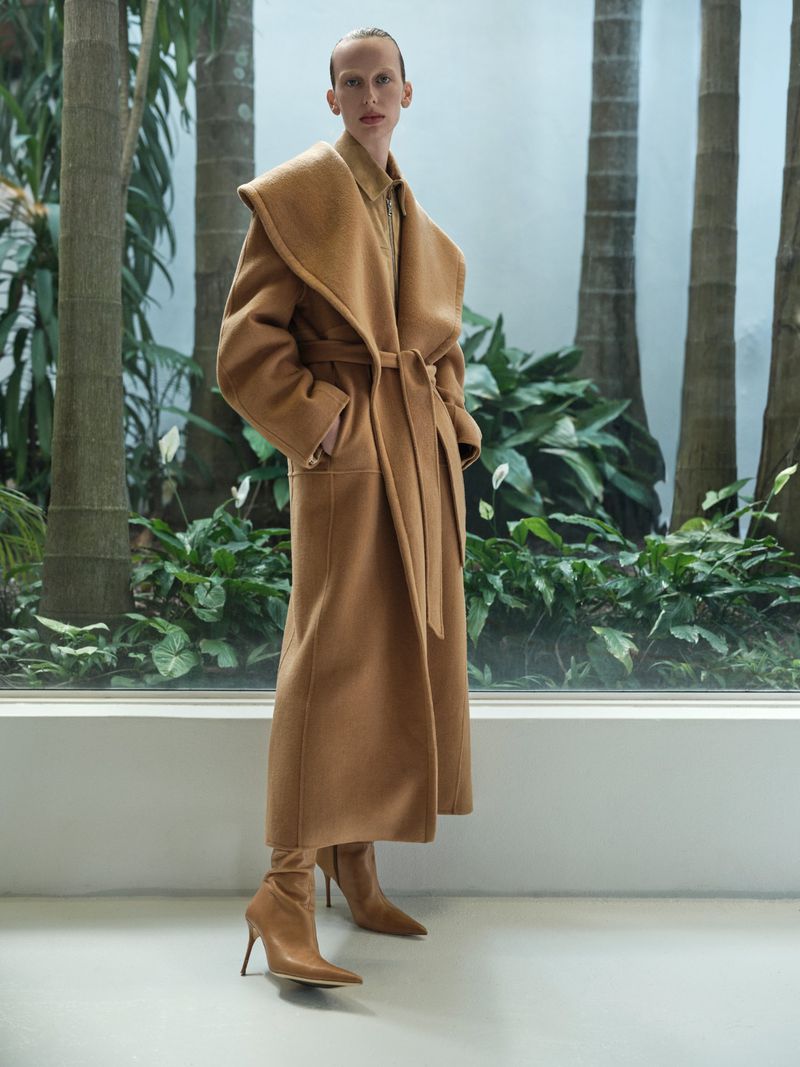 An adult female is posing in a long brown coat, matching boots with heels, in a bright indoor area, surrounded by tropical plants.