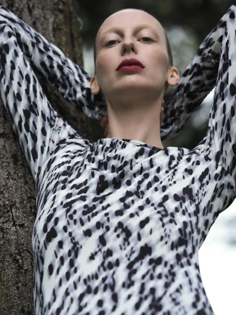 Adult woman in animal print dress poses by tree. Close-up shot. Red lipstick, bare head, fashionable clothing, outdoor, natural light.