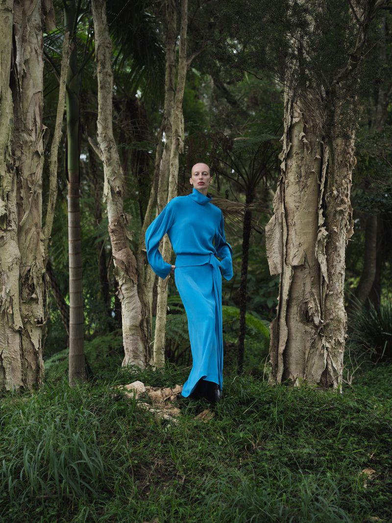 Fashion shot of an adult female in a blue outfit, standing in a forest with tall trees. The shot has a fine art aesthetic.