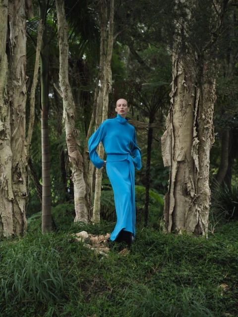 Fashion shot of an adult female in a blue outfit, standing in a forest with tall trees. The shot has a fine art aesthetic.