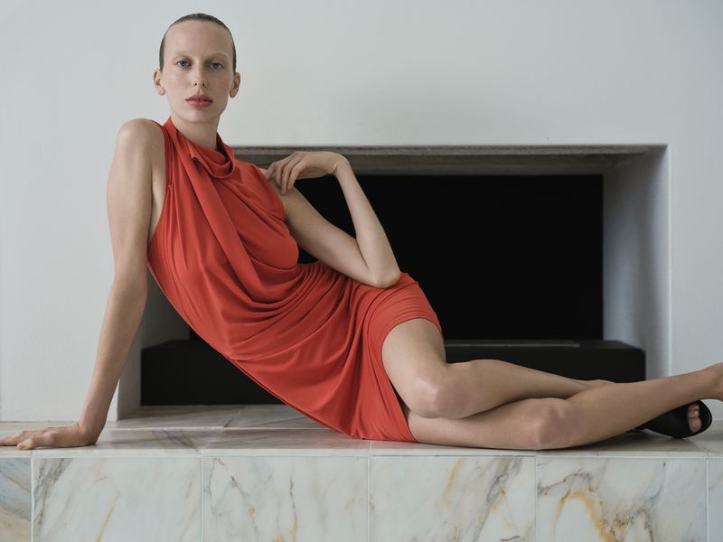A fair-skinned woman in an orange dress poses in a minimalist indoor setting, reclining against a tile structure with a dark backdrop.