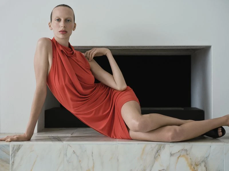 A fair-skinned woman in an orange dress poses in a minimalist indoor setting, reclining against a tile structure with a dark backdrop.