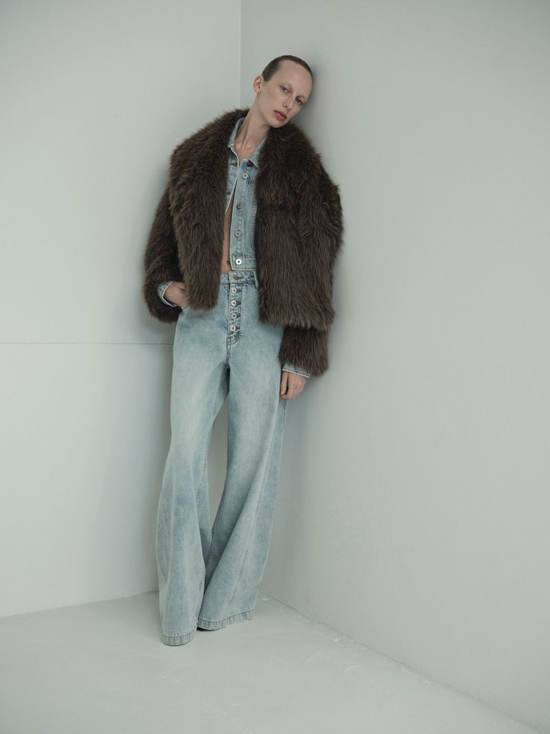 A fair-skinned fashion model wearing a fur coat and denim outfit poses against a neutral corner in a studio setting, showcasing modern style.