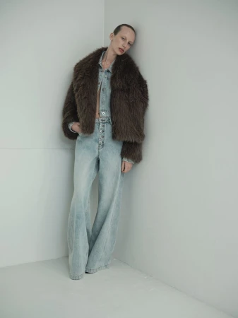 A fair-skinned fashion model wearing a fur coat and denim outfit poses against a neutral corner in a studio setting, showcasing modern style.