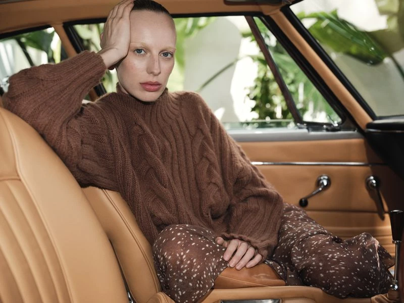 Indoor portrait of a woman with short hair, wearing a brown cable-knit sweater and patterned skirt in a car with tan leather seats.