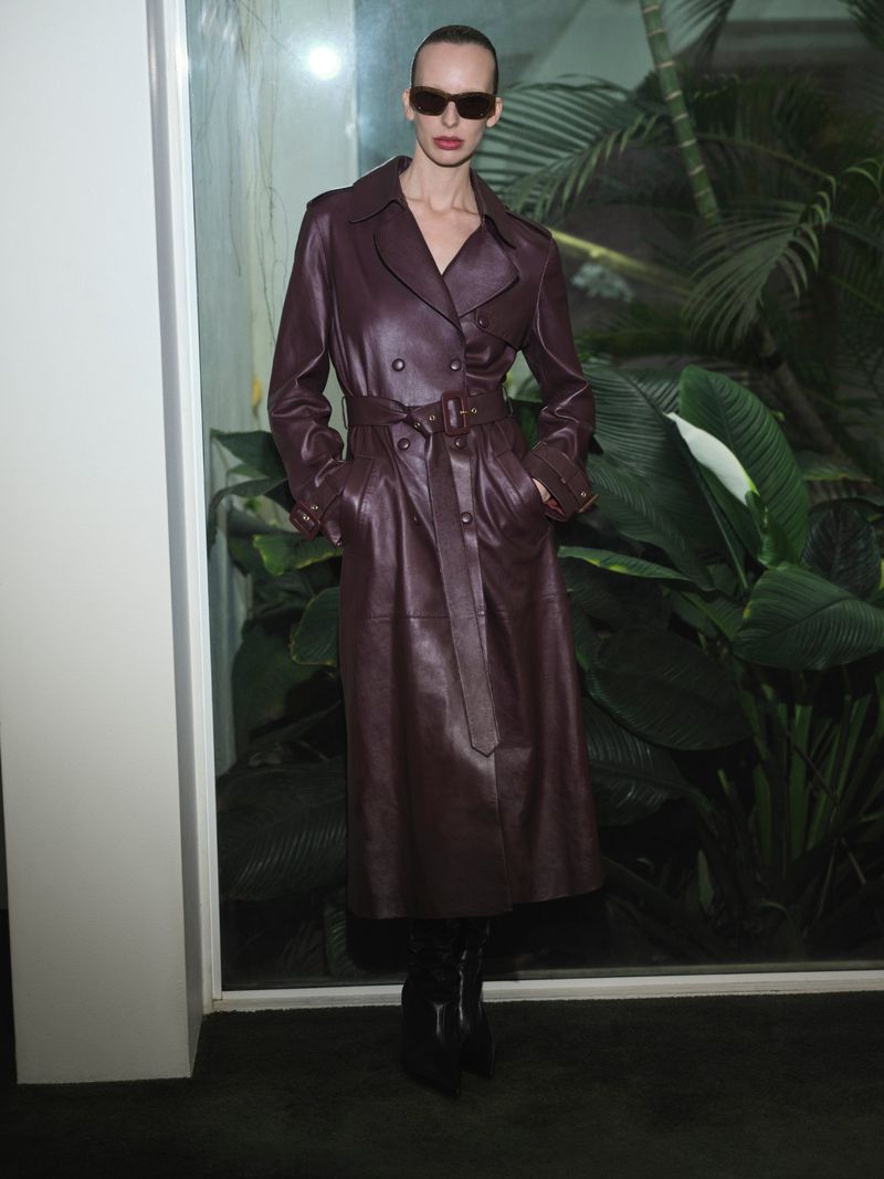 Fashionable woman wearing a dark burgundy leather trench coat with sunglasses poses indoors against green tropical plants.