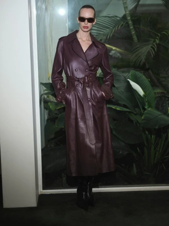 Fashionable woman wearing a dark burgundy leather trench coat with sunglasses poses indoors against green tropical plants.