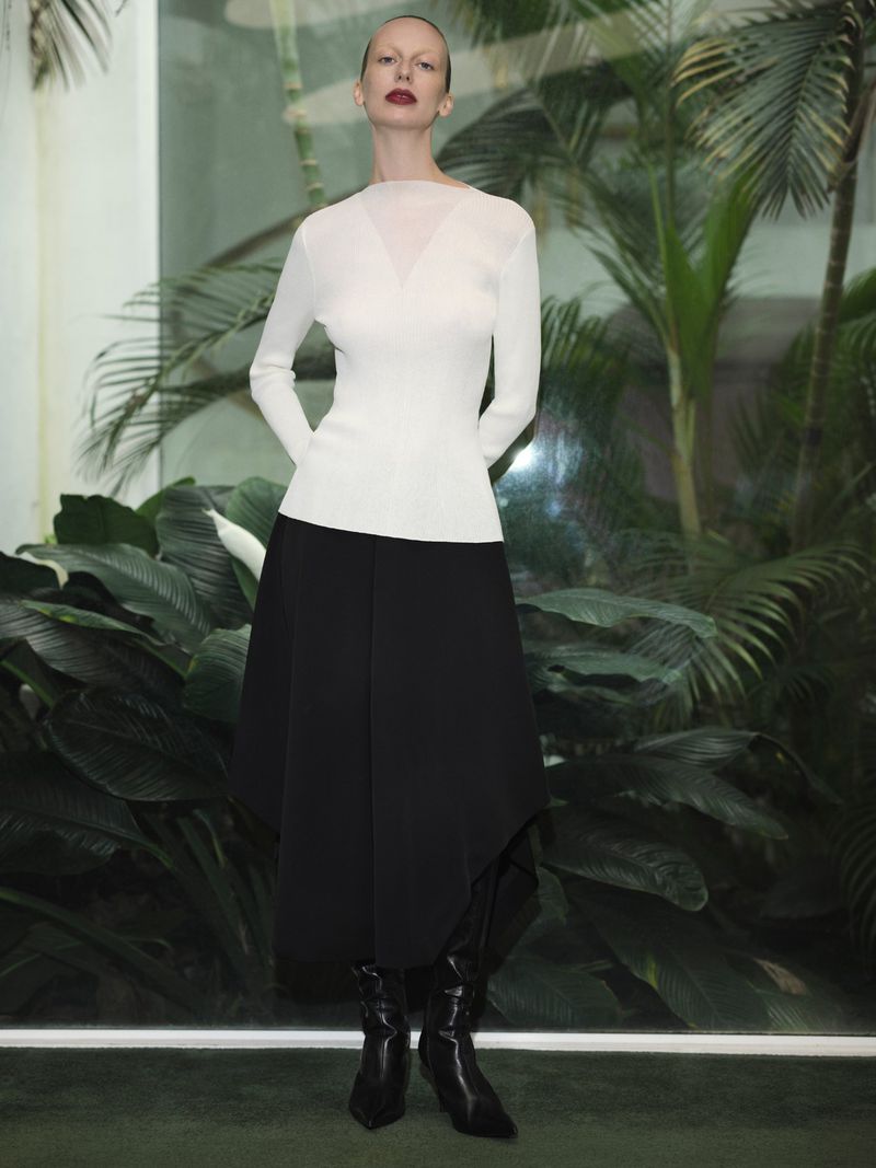 A slender woman with shaved head and red lipstick poses in a white top and black skirt against a backdrop of green tropical foliage.