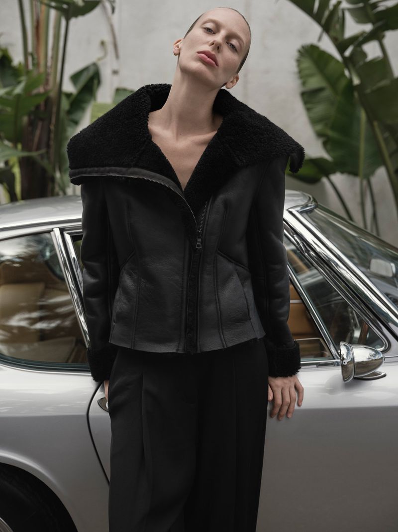 A woman poses in a black shearling coat next to a silver car, set against a backdrop of lush green foliage in an outdoor setting.