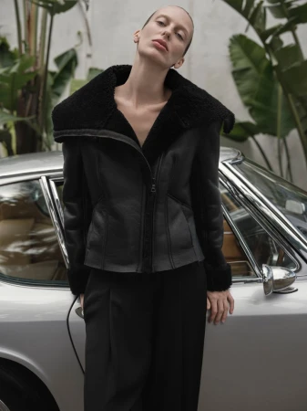 A woman poses in a black shearling coat next to a silver car, set against a backdrop of lush green foliage in an outdoor setting.