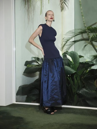 Indoor shot of a slender adult female wearing a navy sleeveless top and navy skirt, posing in front of a leafy background.