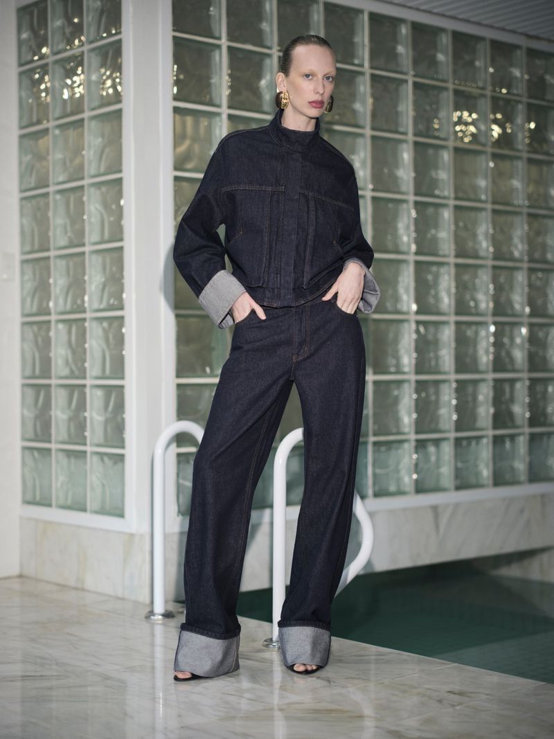 Fashionable adult woman in a dark denim suit with long sleeves poses in front of a tiled indoor pool with marble flooring in studio lighting.