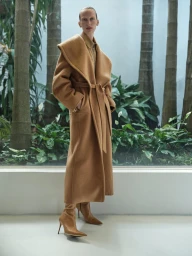 Model Lili Sumner wearing a camel belted coat and boots standing before tropical foliage for Sass & Bide Winter 2025