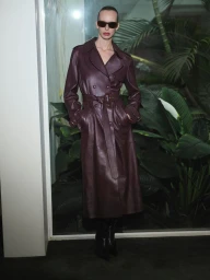 Model Lili Sumner wearing a dark leather trench coat and sunglasses standing before lush tropical foliage.