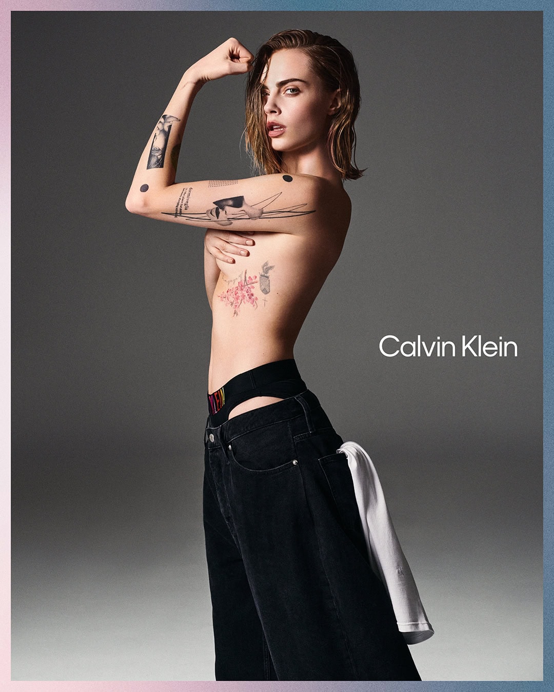 Cara Delevingne poses topless in a studio, wearing loose black jeans and Calvin Klein branded underwear, looking at the camera.