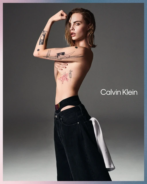 Cara Delevingne poses topless in a studio, wearing loose black jeans and Calvin Klein branded underwear, looking at the camera.