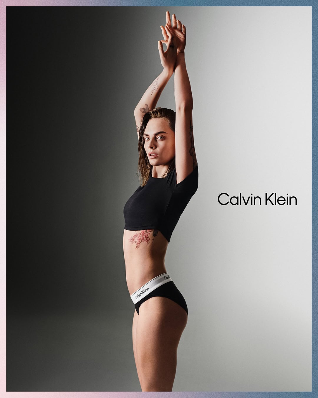A slender, tanned adult female with a tattoo on her torso poses in black underwear and a crop top in a grey gradient studio shot.